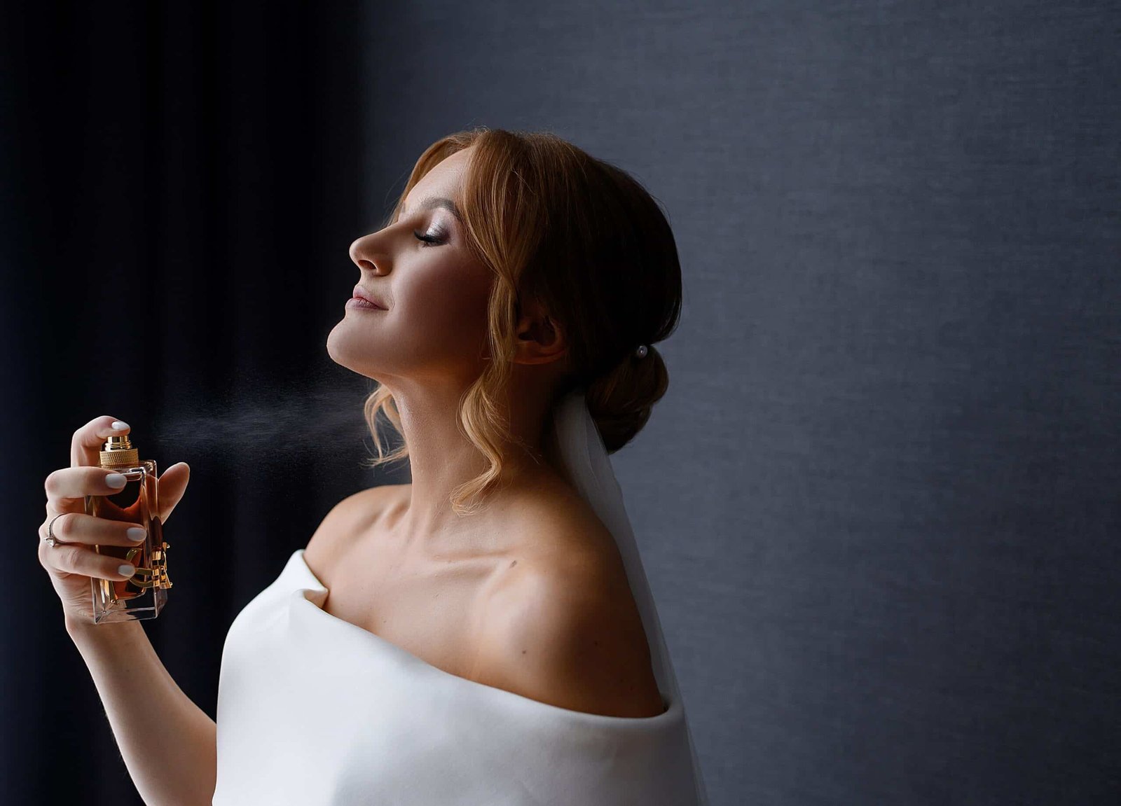 close view sexual bride with makeup hairstyle wearing white silk gown veil spraying perfume her neck enjoying smell against gray background during bridal morning min scaled